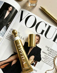 A gold cosmetic tube AND matching cap are arranged on an open Vogue magazine, with a perfume bottle, pen, and jewelry visible in a styled flat-lay setting.