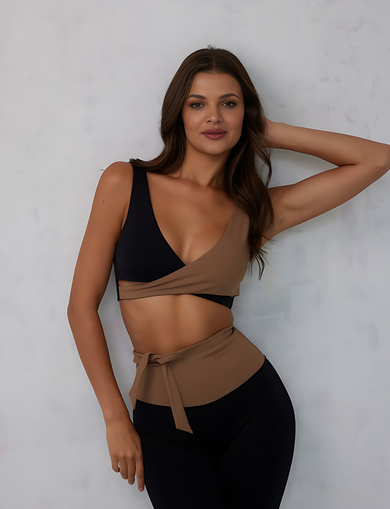 Nubyen Alicante V-Neck Wrap Sports Bra in Biscotti. The bra features a flattering wrap design, deep V-neckline, and smooth fabric for a stylish yet supportive fit. Styled with matching activewear, the look is showcased in a clean, modern setting with soft lighting to emphasize its neutral tone.