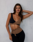 Nubyen Alicante V-Neck Wrap Sports Bra in Biscotti. The bra features a flattering wrap design, deep V-neckline, and smooth fabric for a stylish yet supportive fit. Styled with matching activewear, the look is showcased in a clean, modern setting with soft lighting to emphasize its neutral tone.