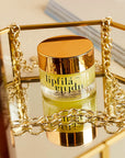 A jar of lipfilá lip balm with a shiny gold lid and clear container showing yellow-green balm inside, styled on a reflective gold tray with chain accents.