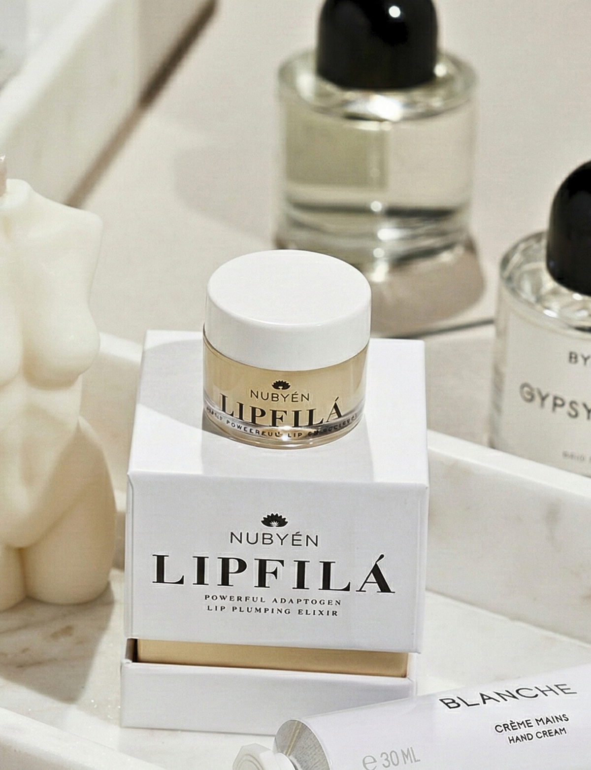 Nubyén Lipfilá lip plumping elixir shown with its white outer box, a gold inner tray, and the open jar placed on top. BEST SELLER