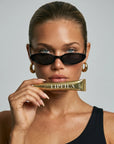 A close-up of a model wearing black cat-eye sunglasses and gold hoop earrings, holding a gold tube of Lipfila lip gloss near her mouth against a light gray background.