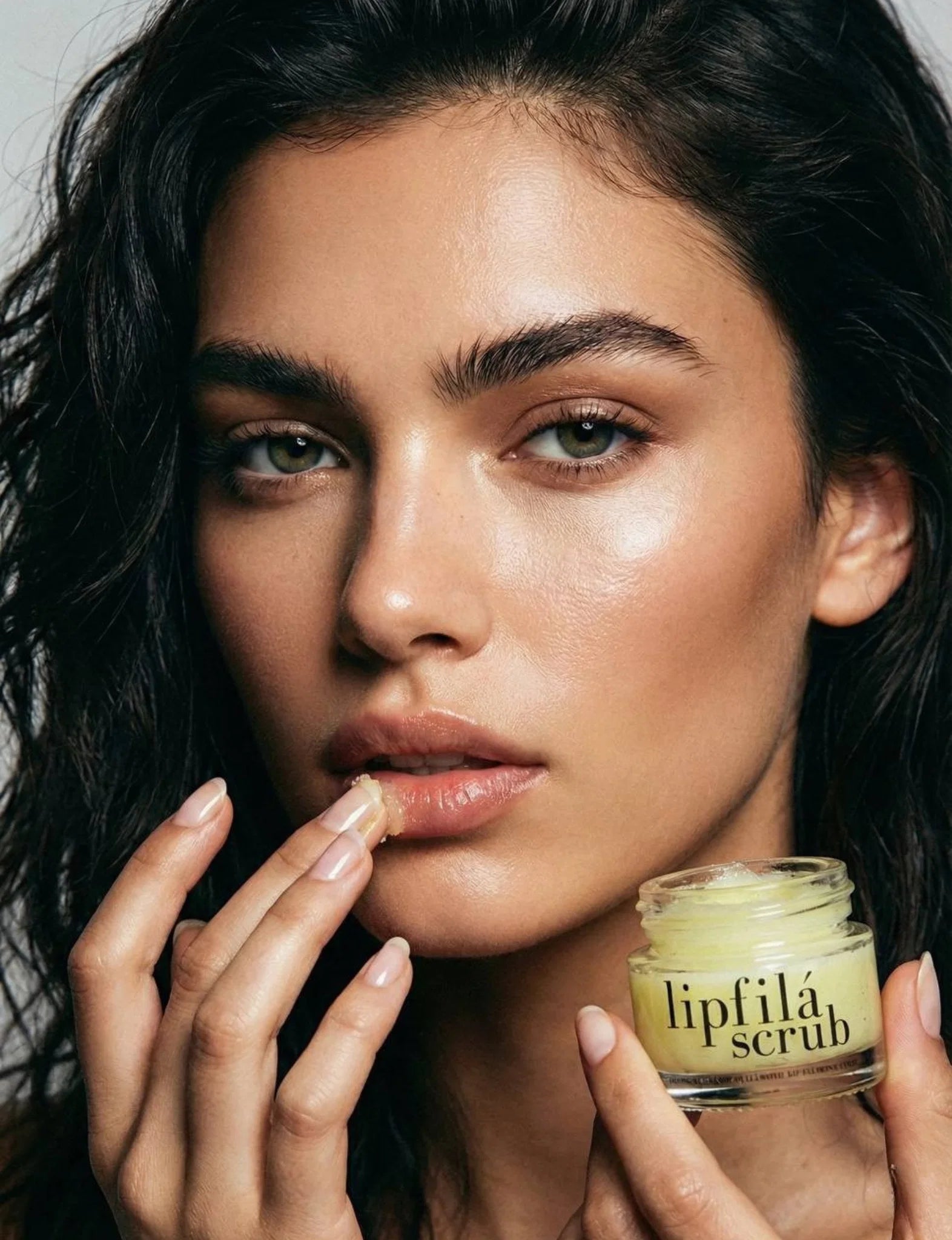 Close-up of a woman holding a small clear jar of lipfilă scrub and touching her lips, with the product jar visible in the lower right against a soft light gray background.